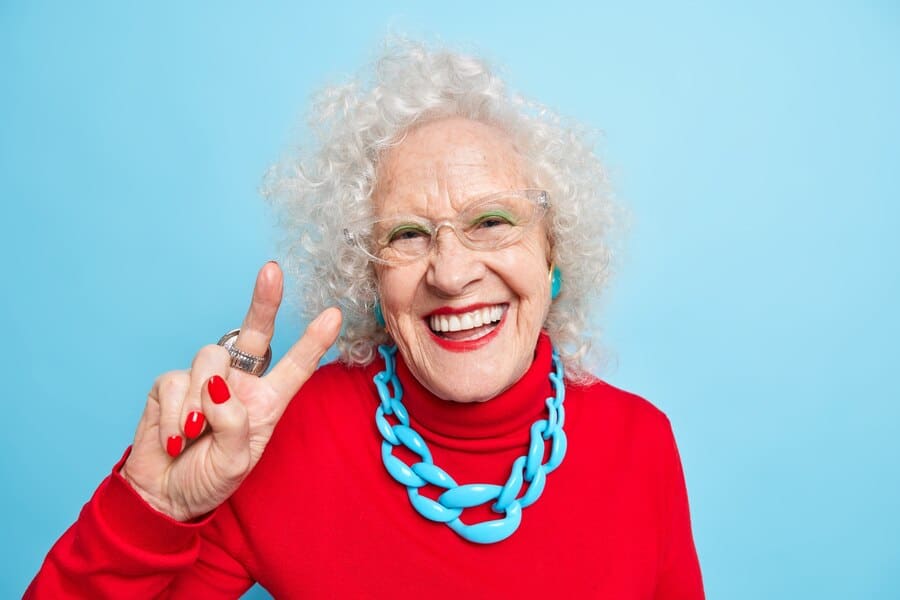portrait cheerful nice looking elderly woman smiles happily makes peace gesture shows v sign dressed red jumper with necklace expresses positive emotions 273609 50092